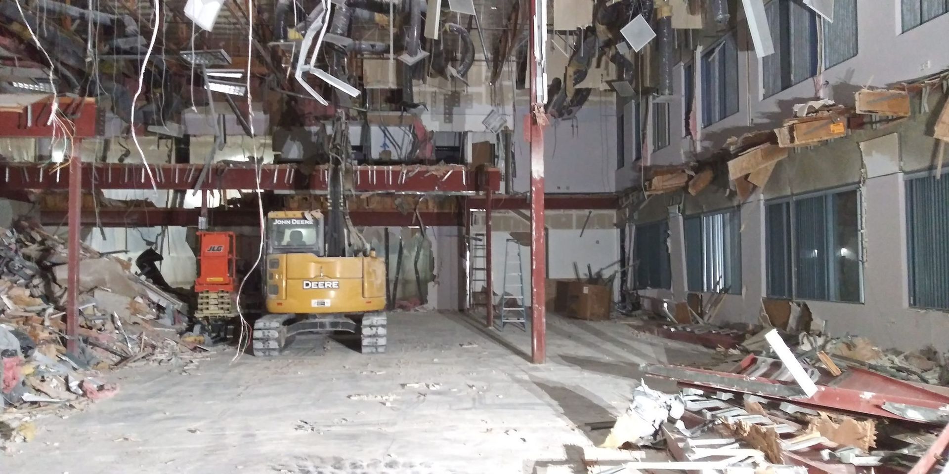 Damaged warehouse interior with debris, a yellow excavator, and broken ceiling panels everywhere