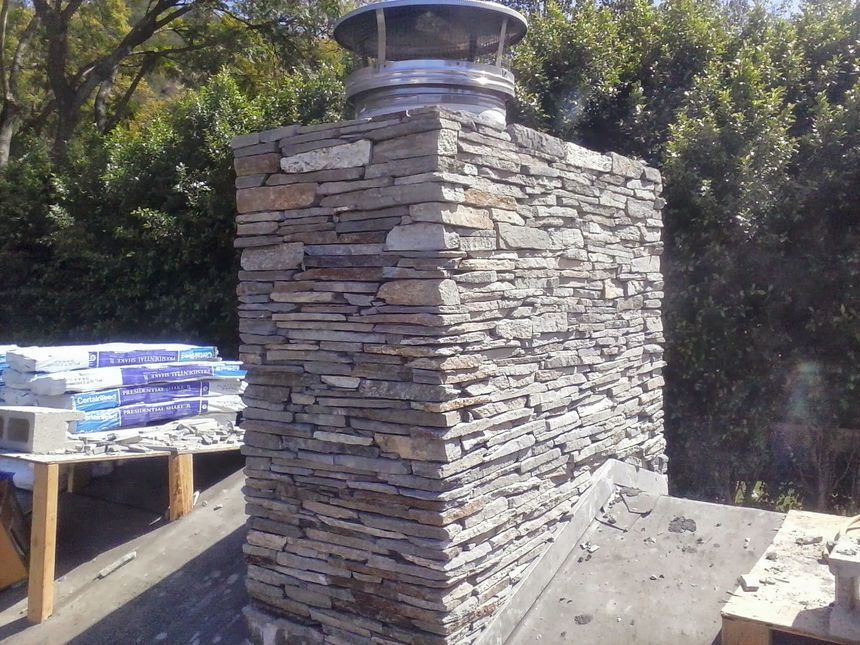 Stacked stone outdoor fireplace chimney beside a wooden table in a sunny yard.