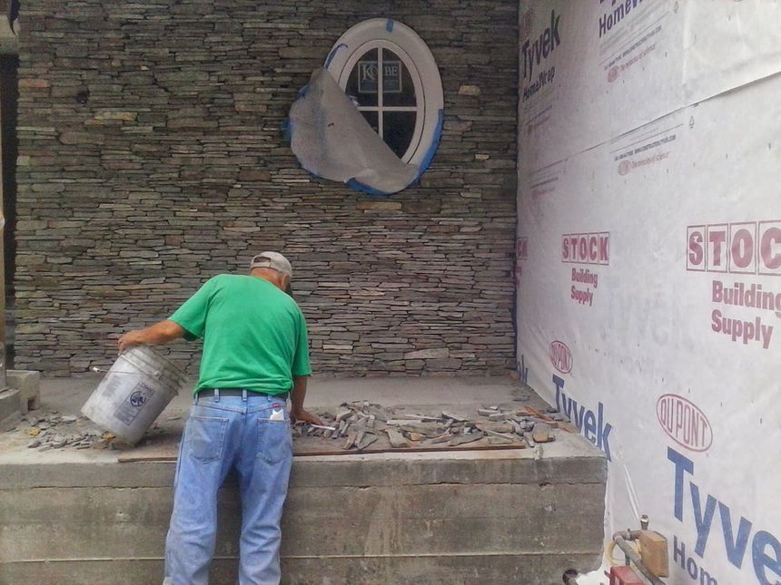 Worker in green shirt pours a bucket of rubble at a construction site with stone wall and Tyvek wrap.