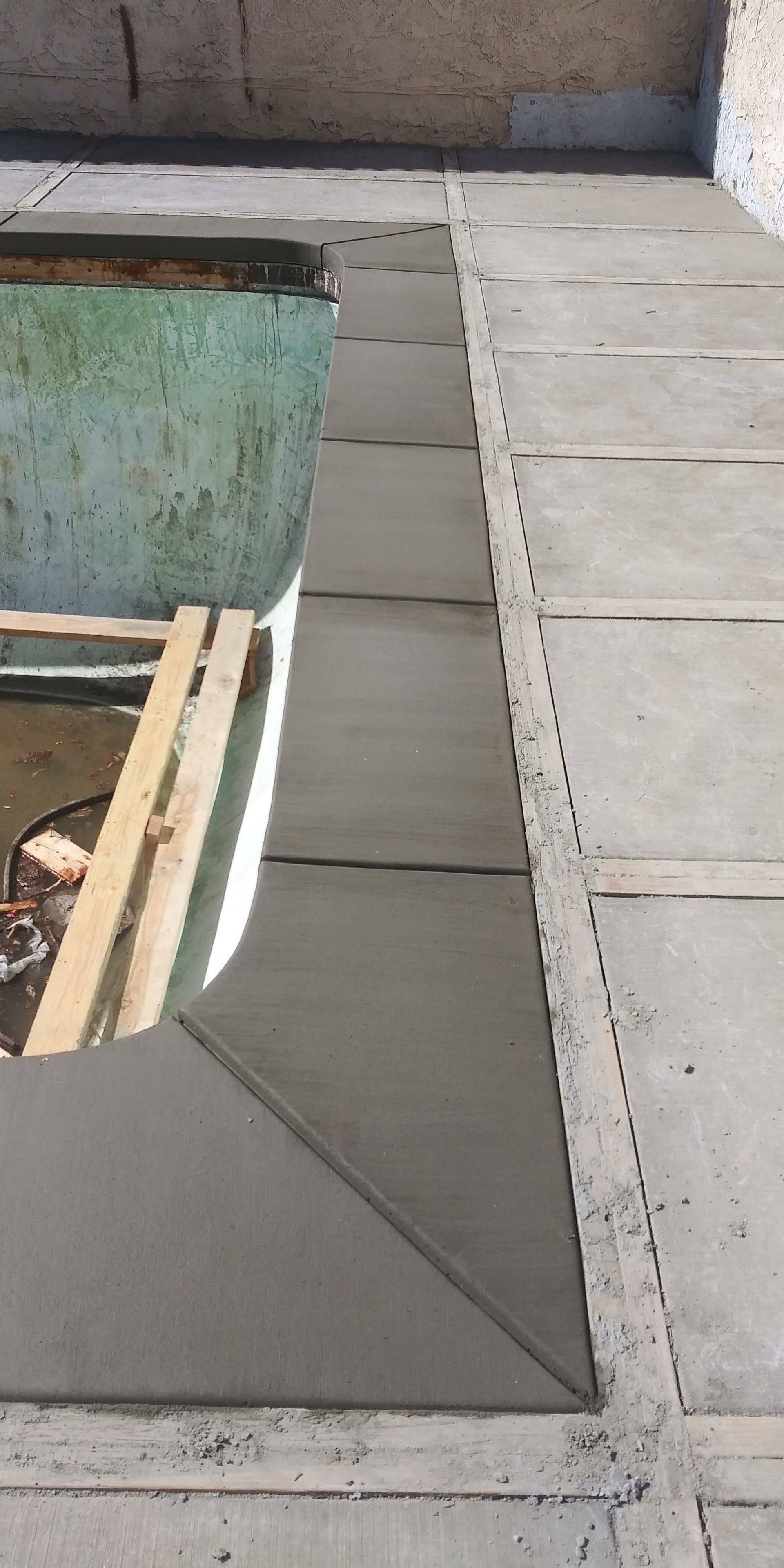 Concrete patio beside a pool, with a green pool wall and wooden planks in the foreground.