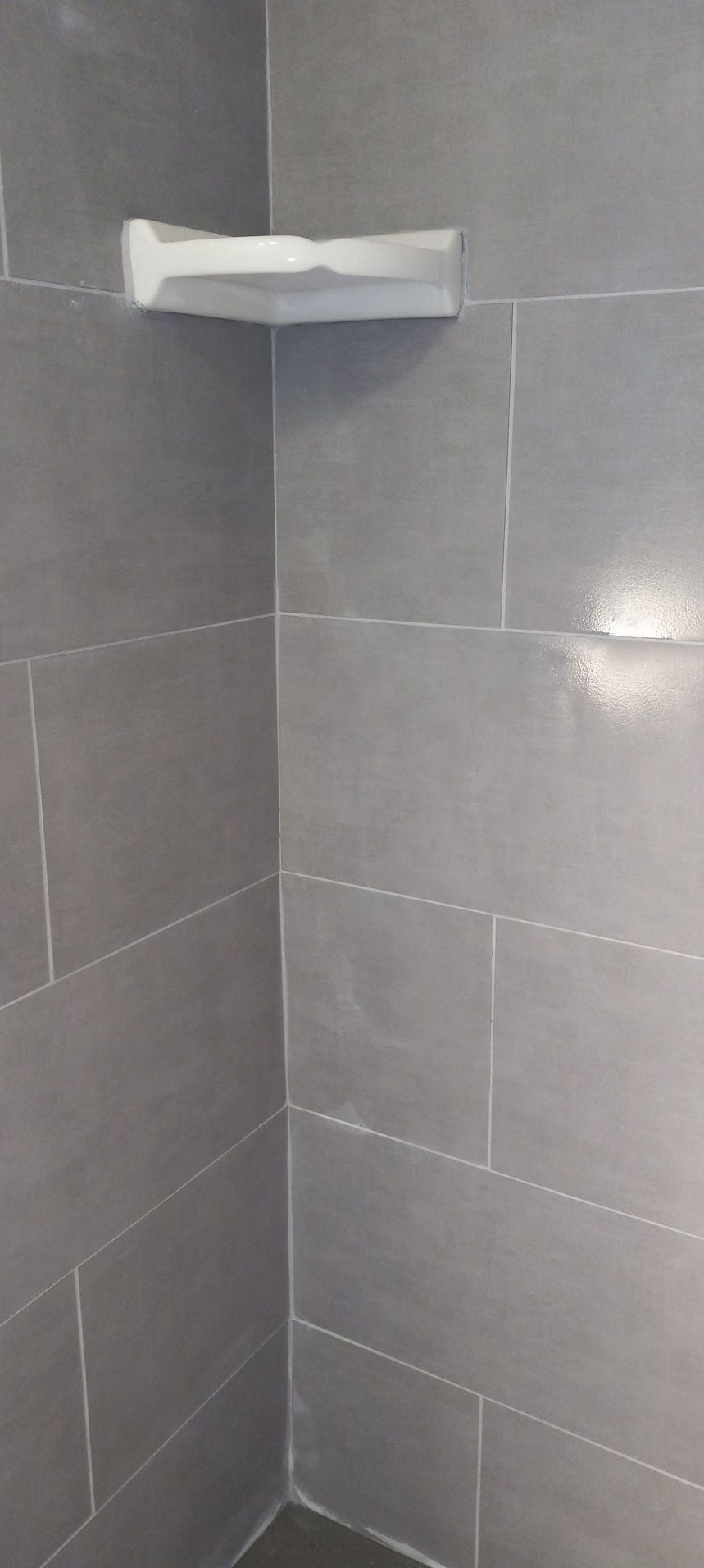 Gray tiled shower corner with a white corner shelf mounted near the top.