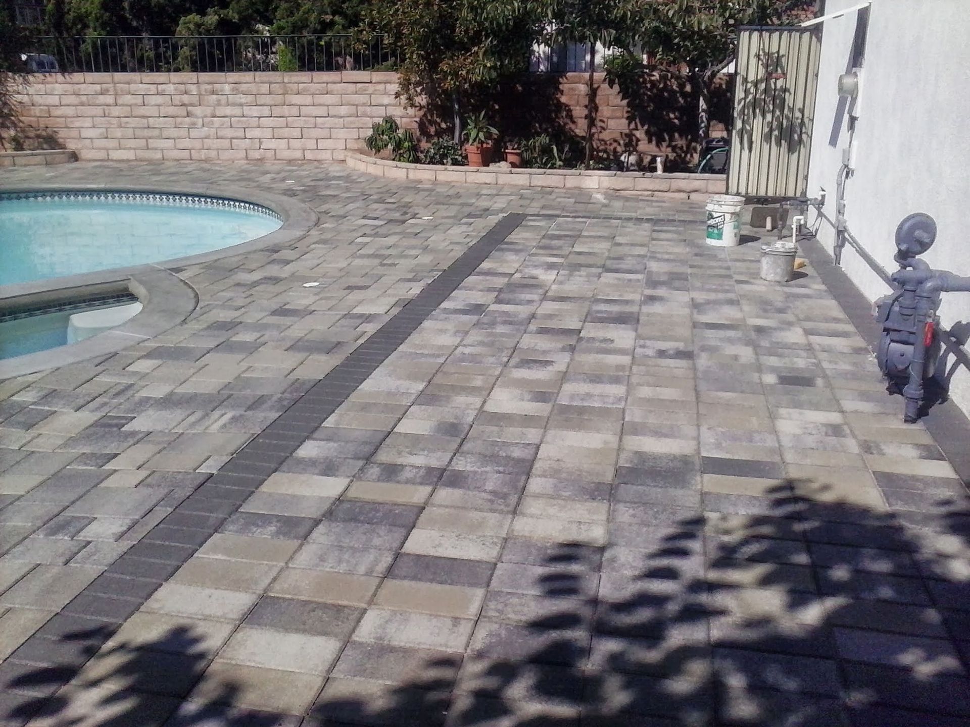Poolside patio with gray stone pavers, a small round pool on the left, and shadows from nearby trees.
