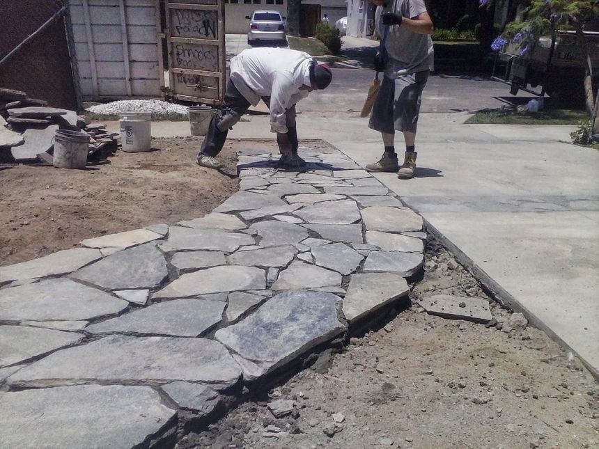 Two workers fitting large stone slabs into a walkway beside a concrete path outdoors