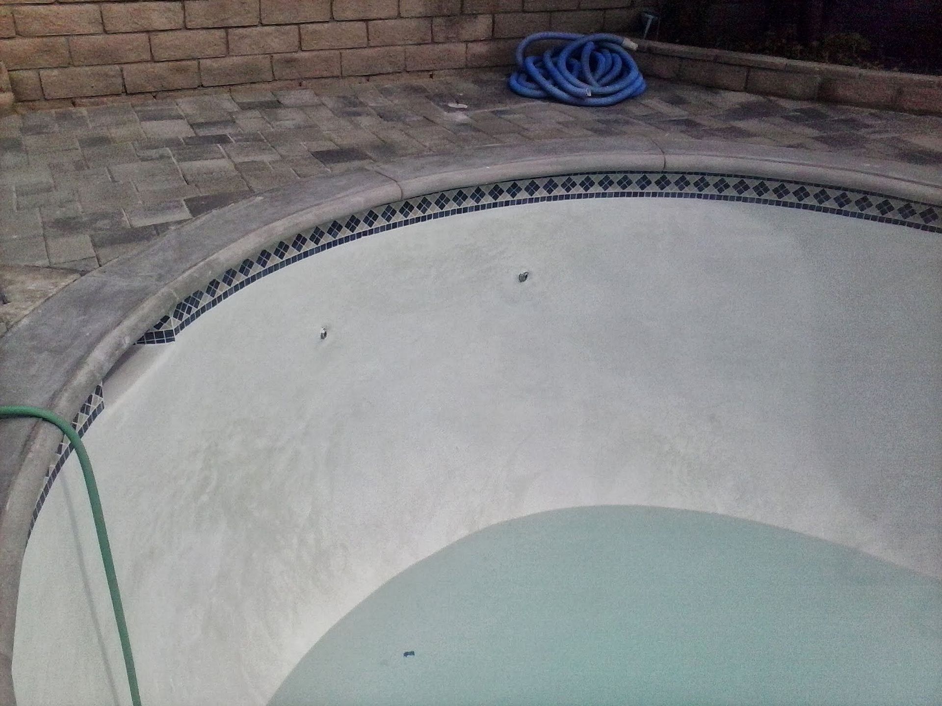Empty swimming pool with light blue water and a coiled hose on the deck beside a brick wall