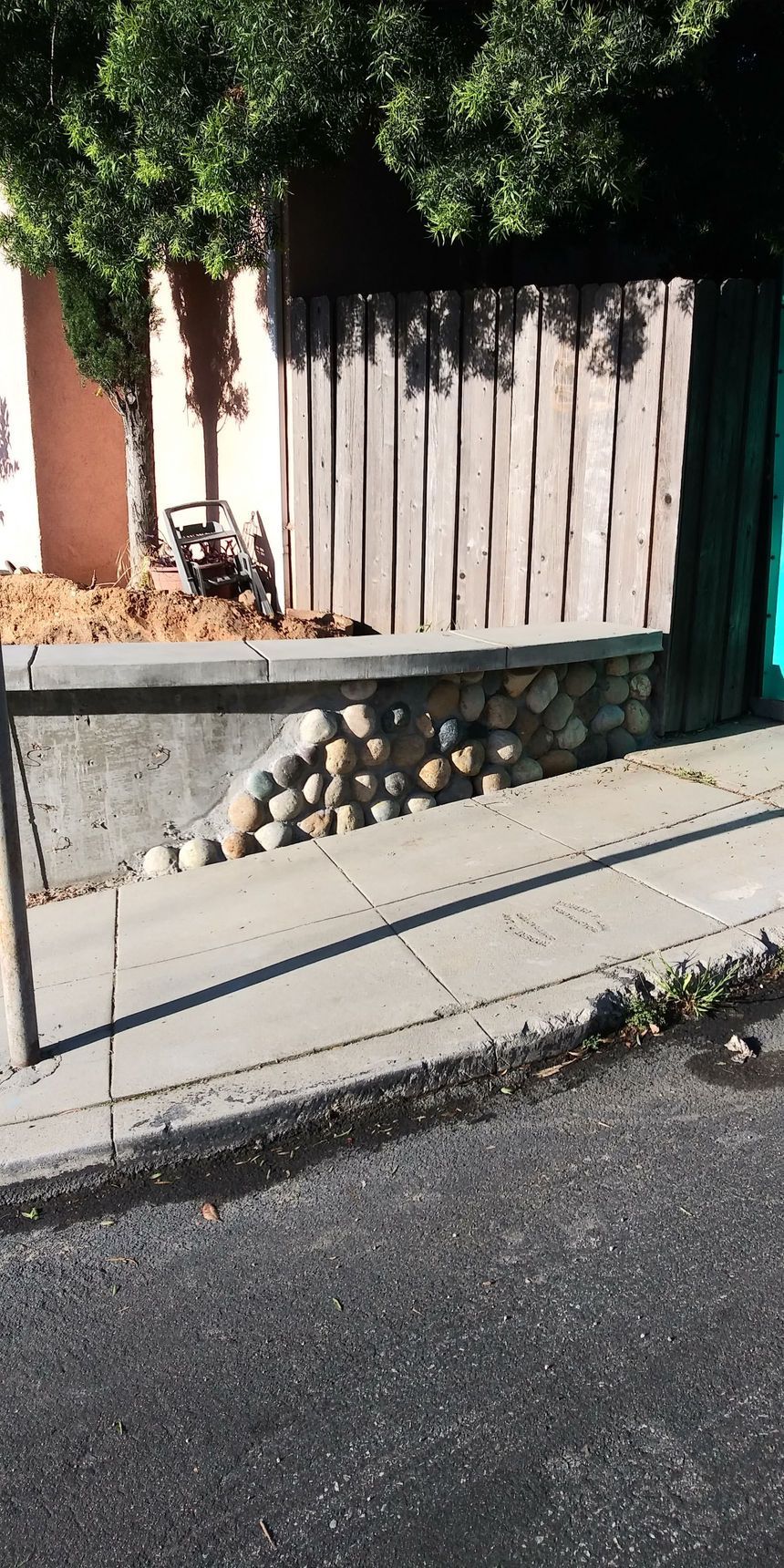 Residential sidewalk with a low stone wall, wooden fence, and greenery along the street