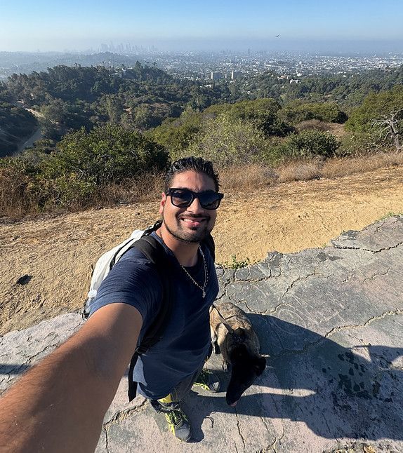 Selfie on a sunny hilltop with city skyline in the background, wearing sunglasses and a backpack.