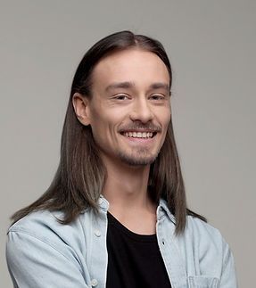 Smiling person with long brown hair wearing a light blue shirt over a black T-shirt against a gray background