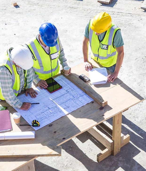 Construction workers in hard hats review blueprints at a table on a job site.