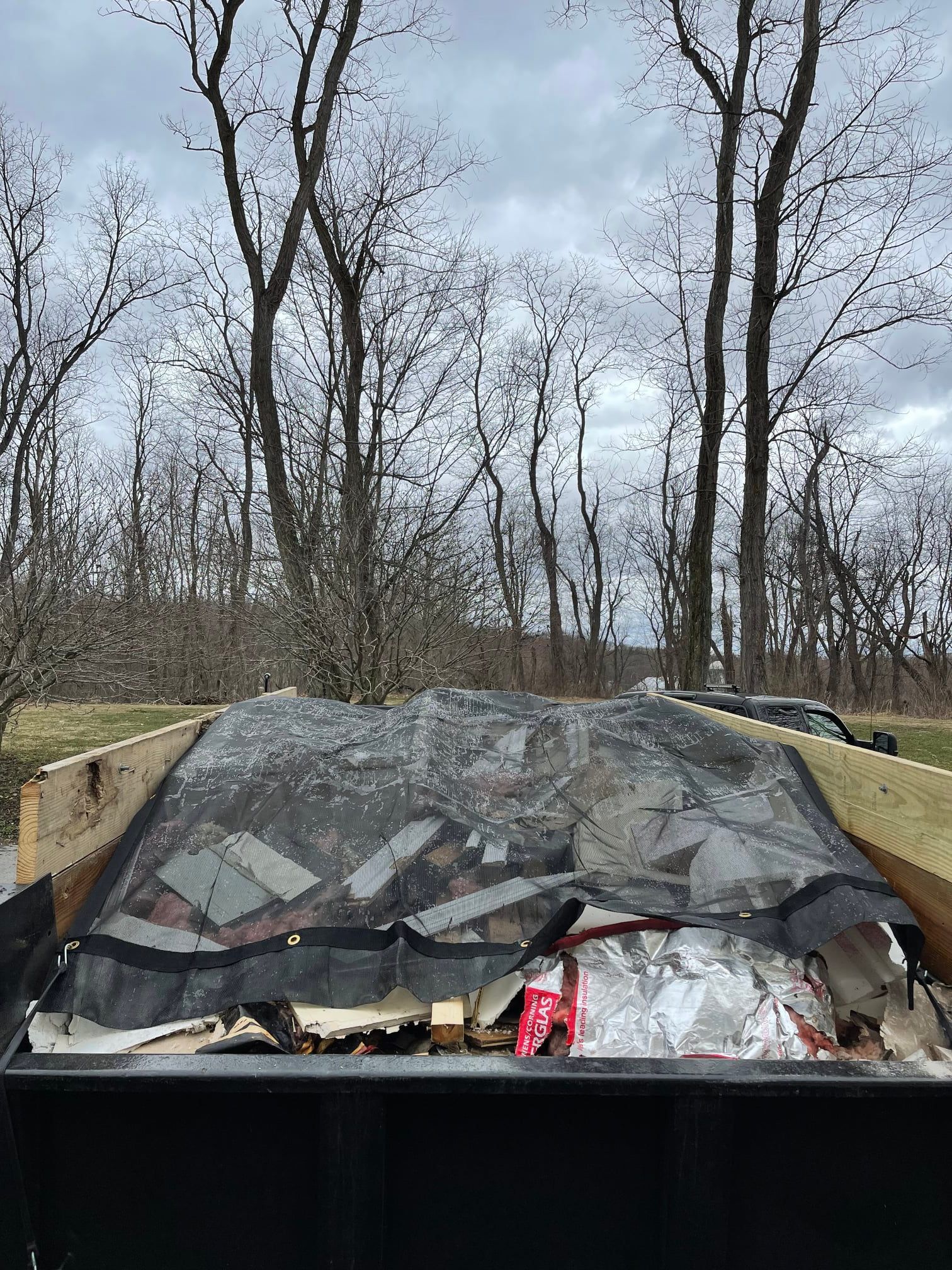 Crushed car windshield with debris inside, parked under bare trees on a cloudy day.