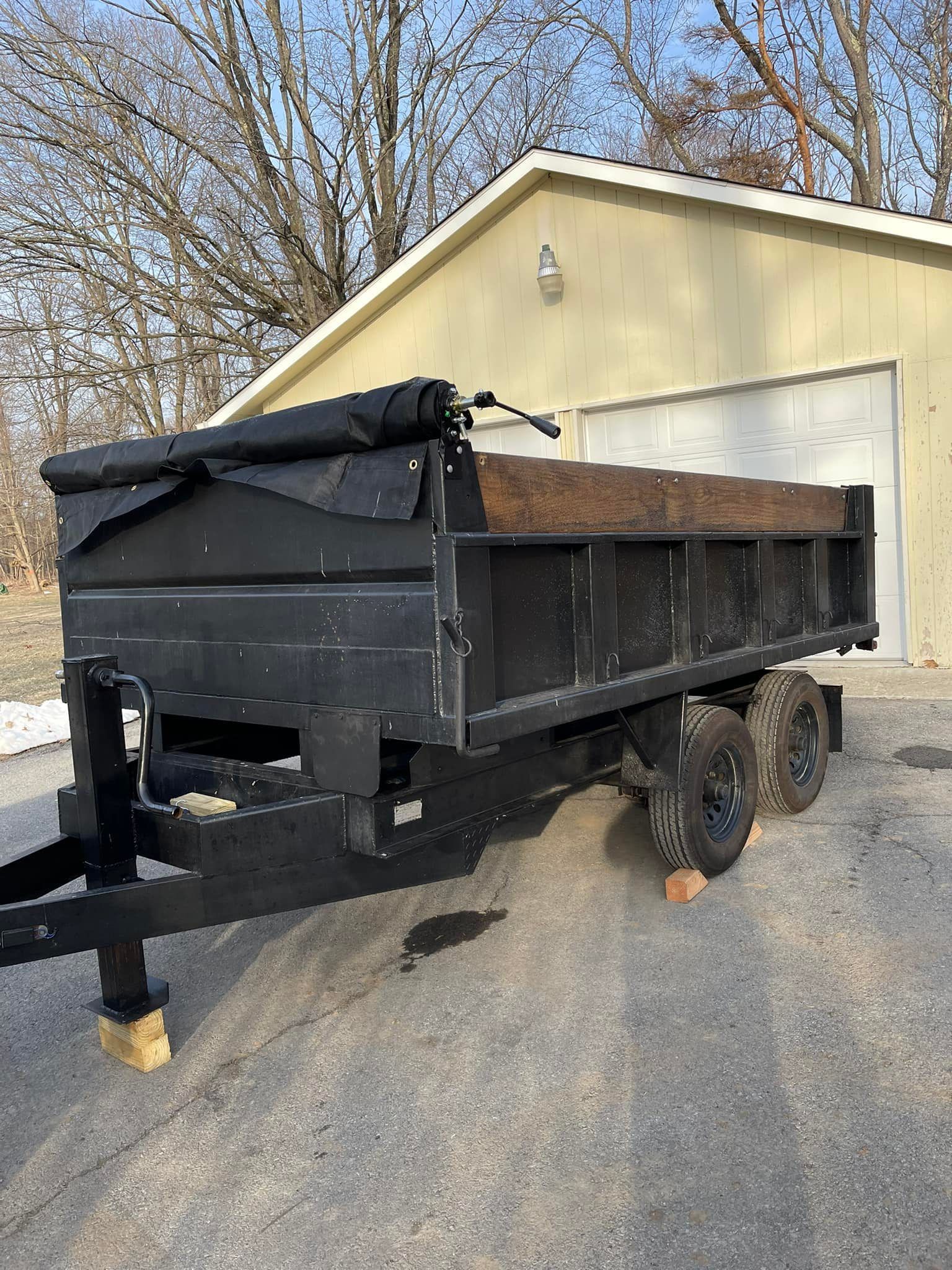 Black dump trailer with tarp parked on gravel in front of a beige garage and trees