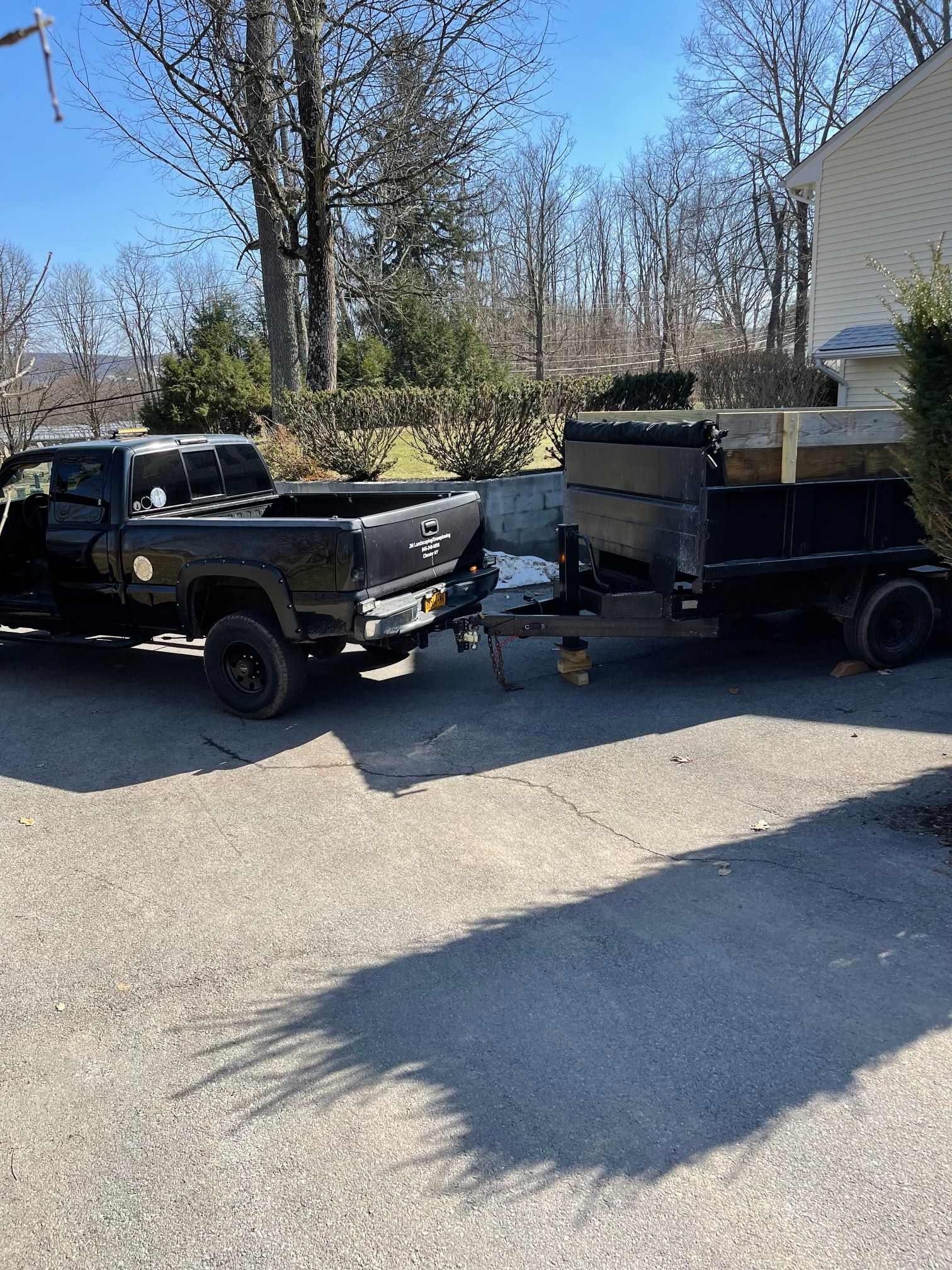Black pickup and utility trailer parked in a driveway beside a house, with trees in the background.