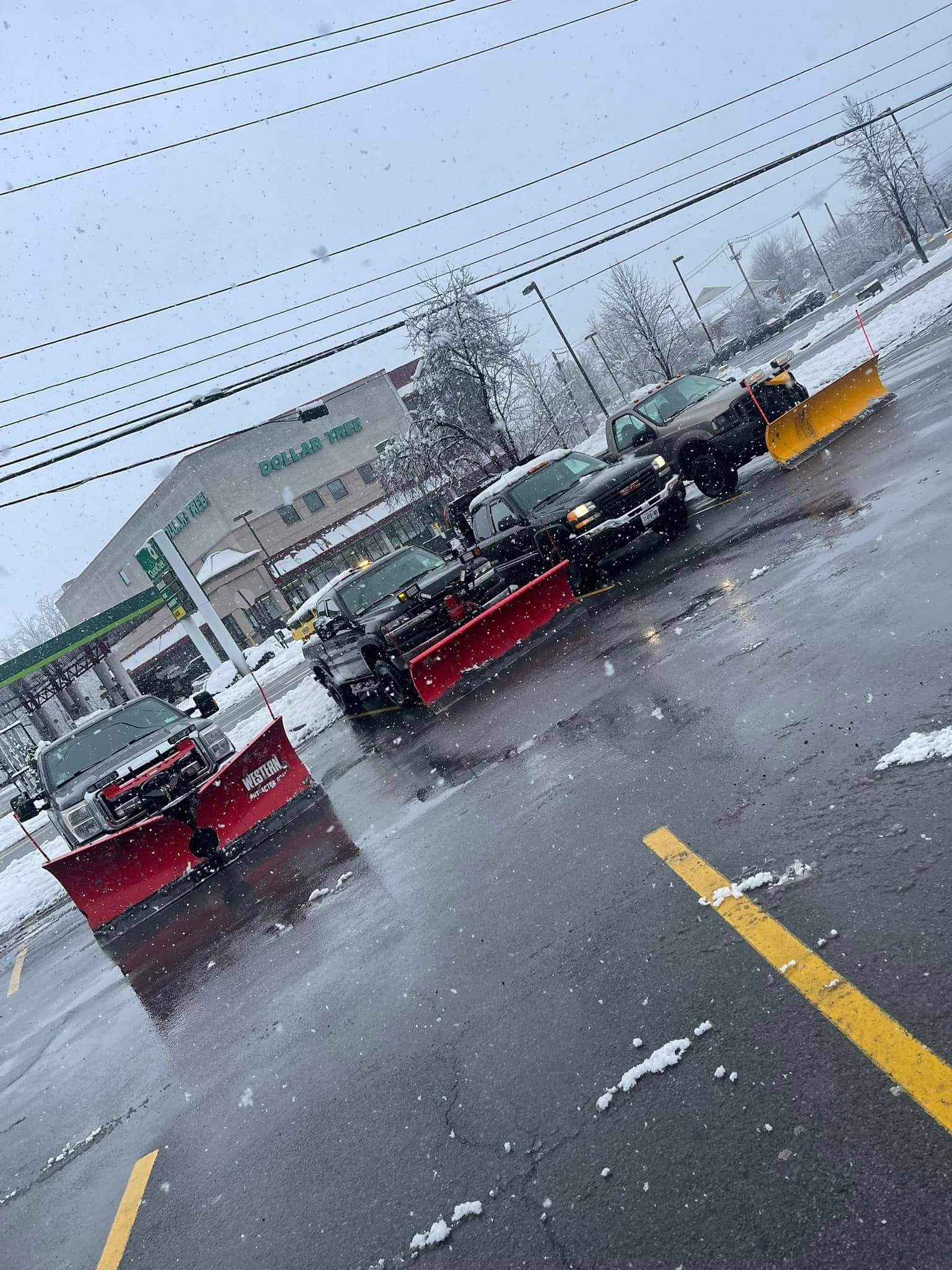 Snowy parking lot with several snowplows clearing slushy pavement under an overcast sky