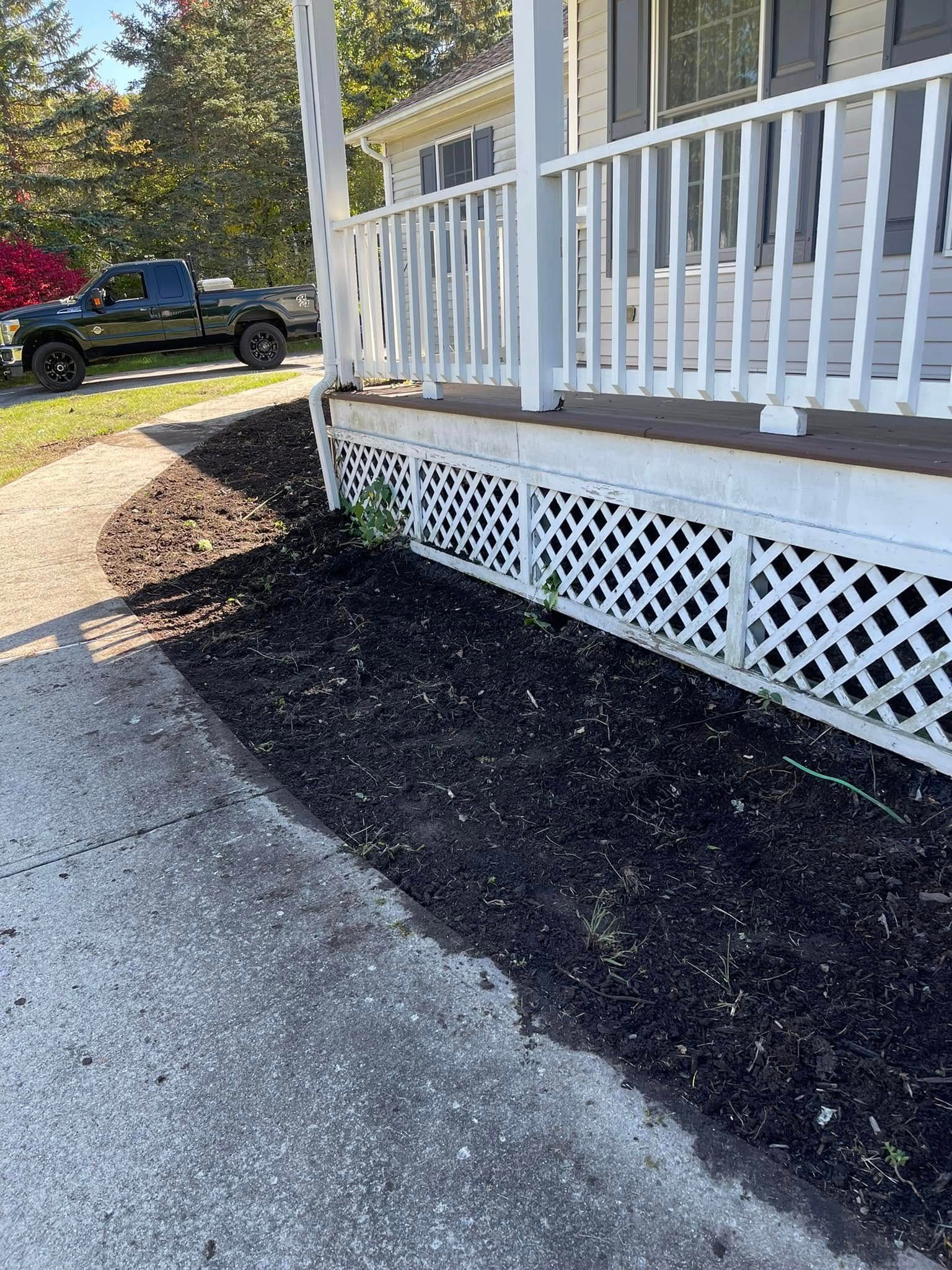 White porch with lattice skirting beside a gravel driveway and dark mulch bed, with a truck in the background