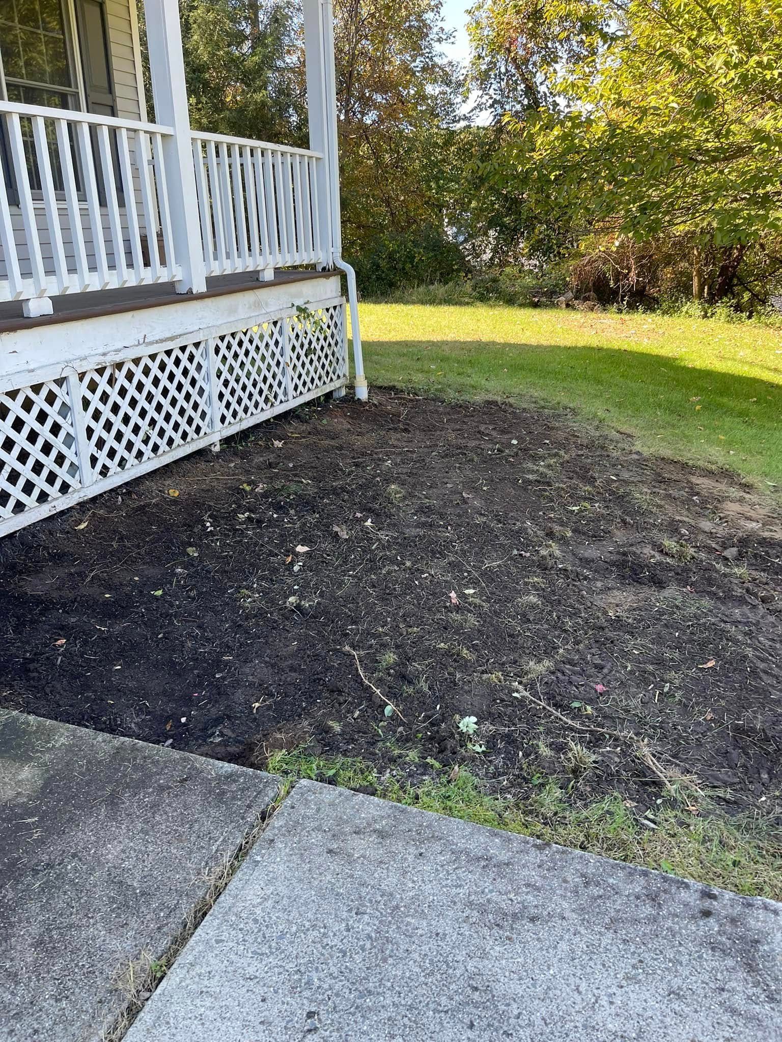 Mulched garden bed beside a white porch and concrete walkway, with grass and shrubs in the background