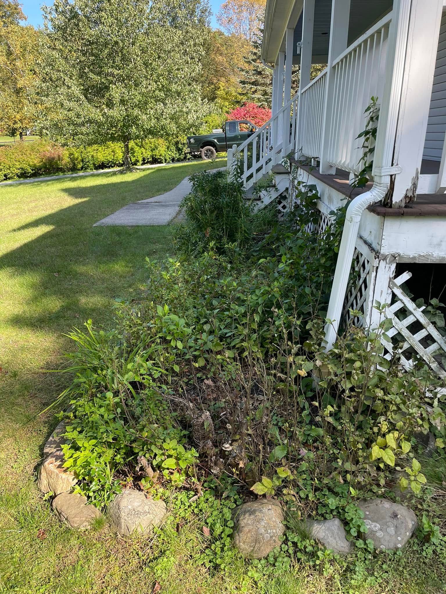 Front yard garden bed beside a white porch, with green shrubs, rocks, and a lawn walkway.