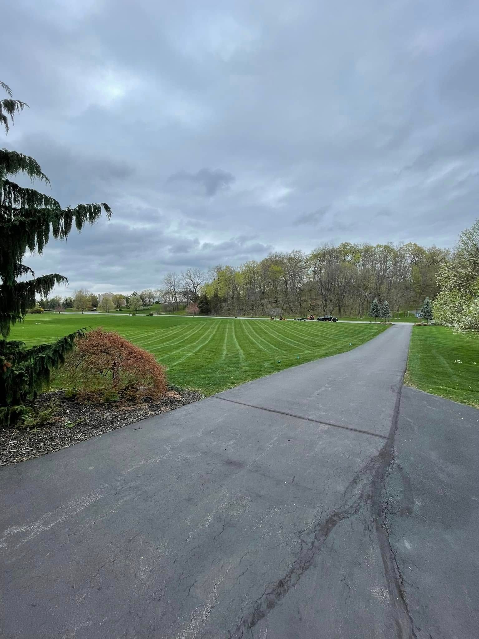 Paved path beside a grassy field and trees under a cloudy sky