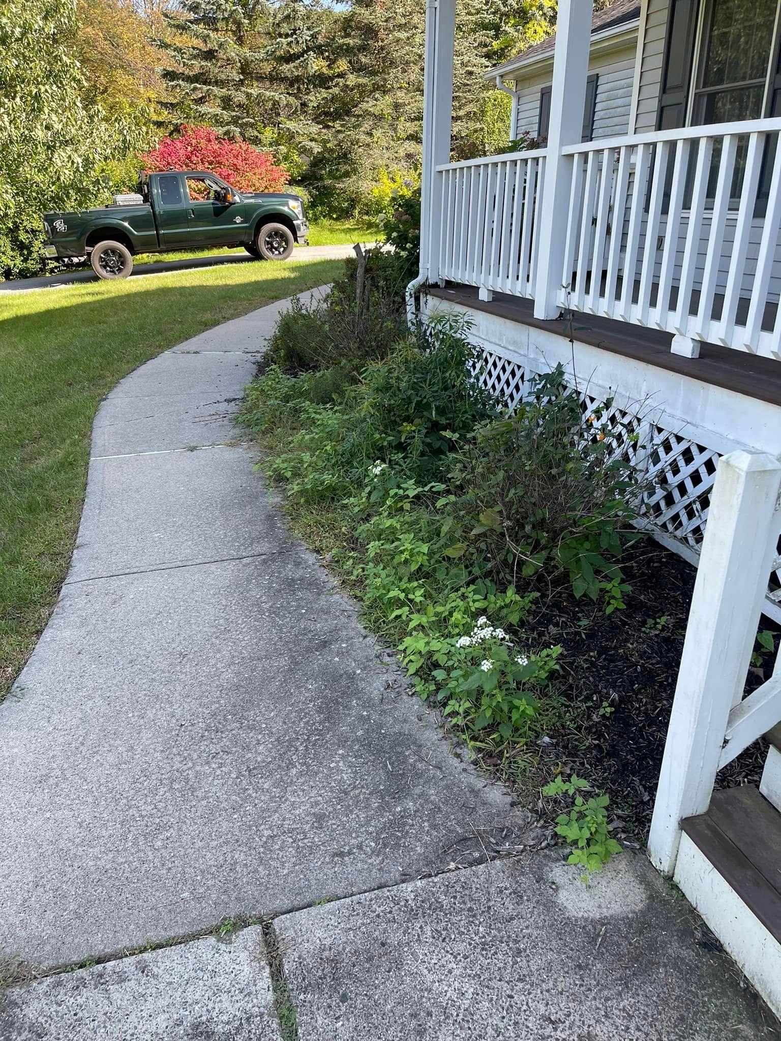 Curved concrete sidewalk beside white porch railing and shrubs, with a pickup truck parked on the lawn.