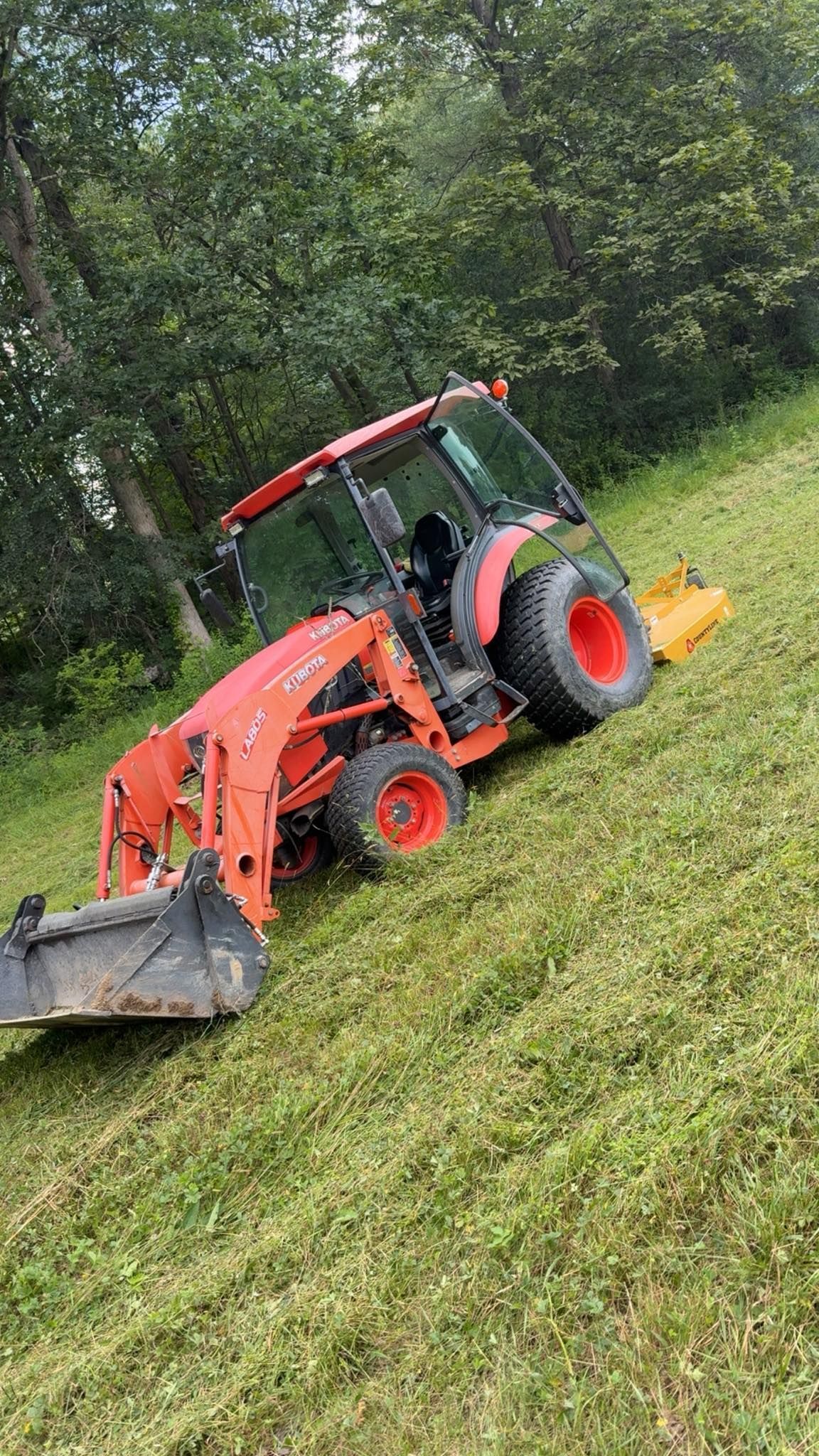 Orange tractor mowing grassy hillside with front loader and rear attachment