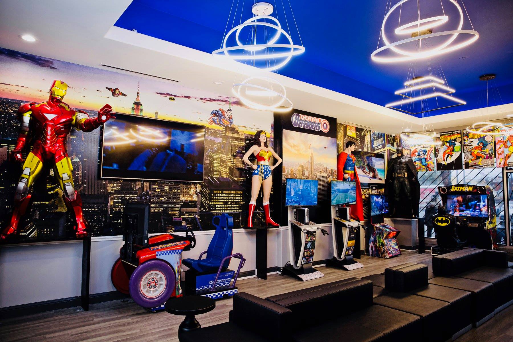 Bright arcade with superhero displays, colorful game cabinets, and neon ring lights under a blue ceiling