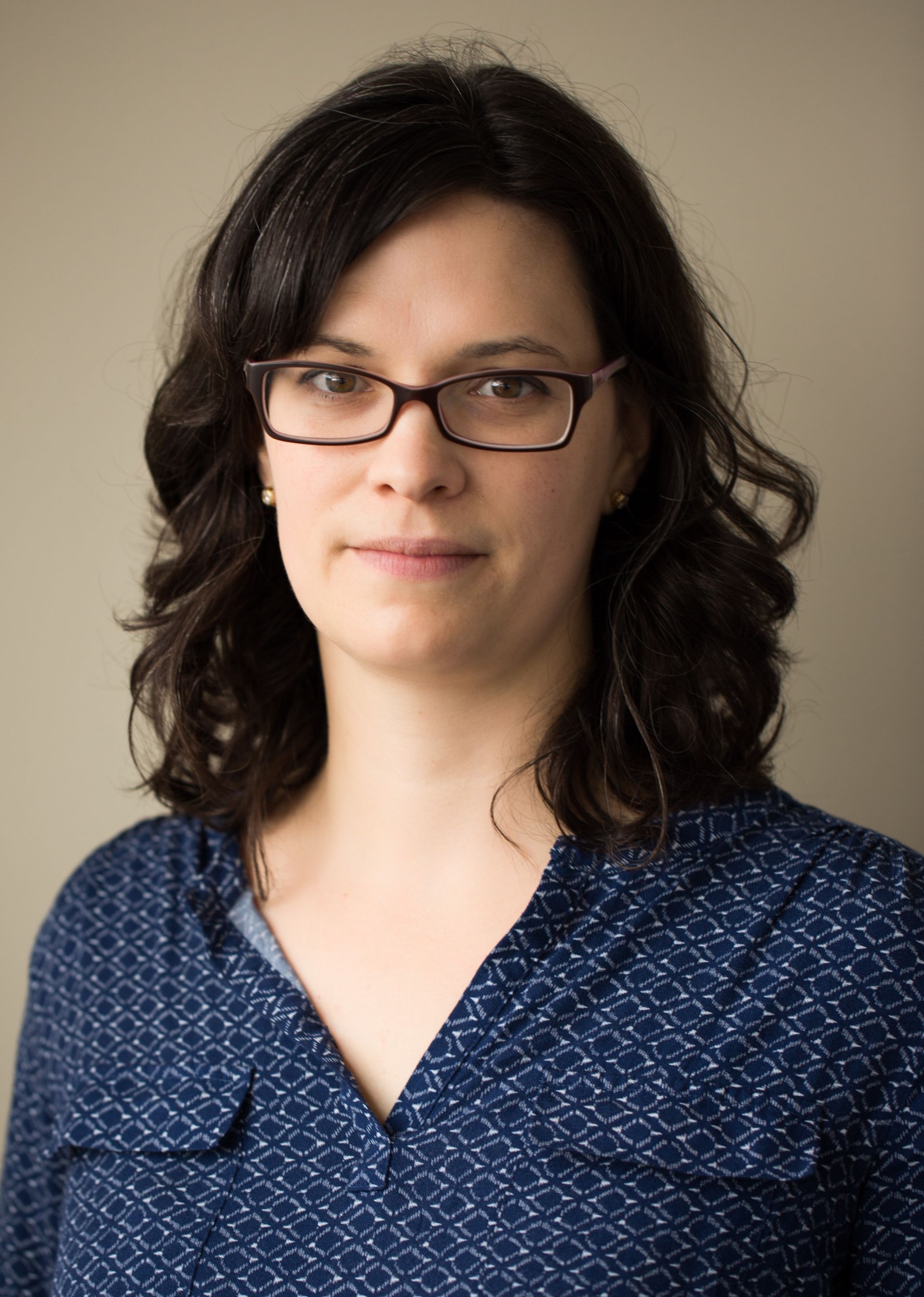 A woman wearing glasses and a blue shirt is standing in front of a wall.