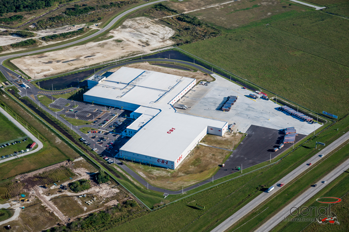 Aerial view of a large industrial building with a parking lot, trucks, and surrounding green fields.