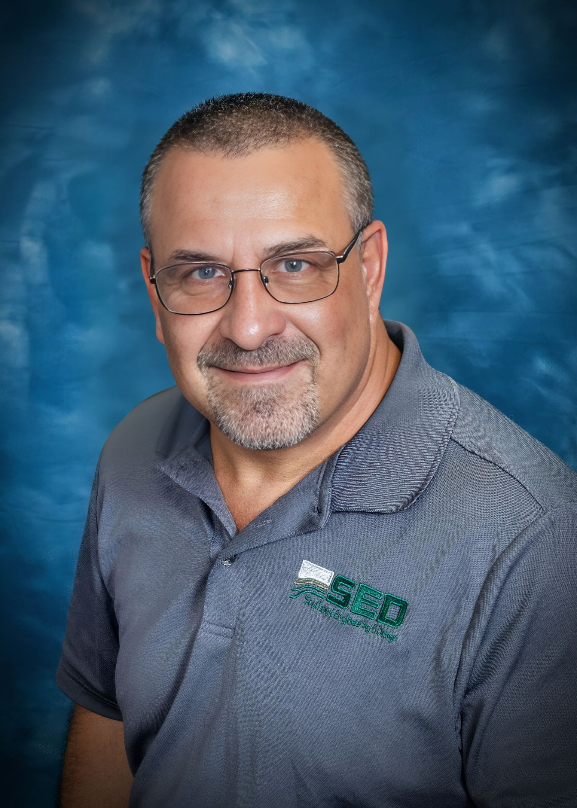 Man with glasses, gray polo shirt, smiling in front of a blue backdrop.