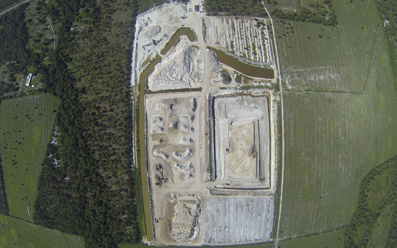 Aerial view of a construction site surrounded by trees and fields, showing earthworks and structures.