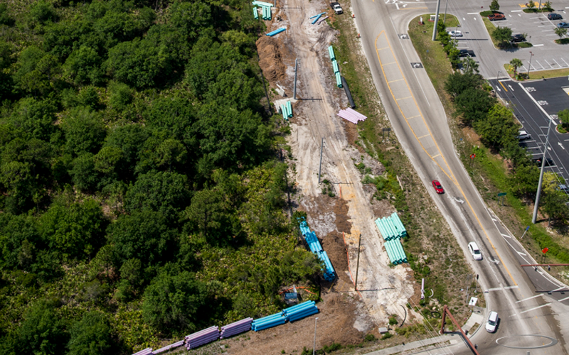Aerial view of road construction next to green trees and a road with cars. Construction materials are present.
