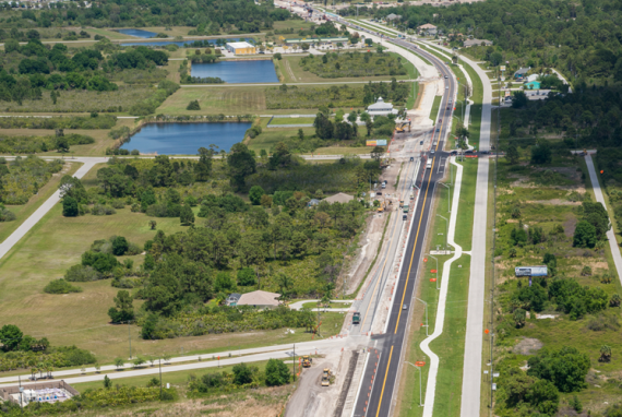 Aerial view of a multi-lane highway under construction with wetlands and buildings in the background.
