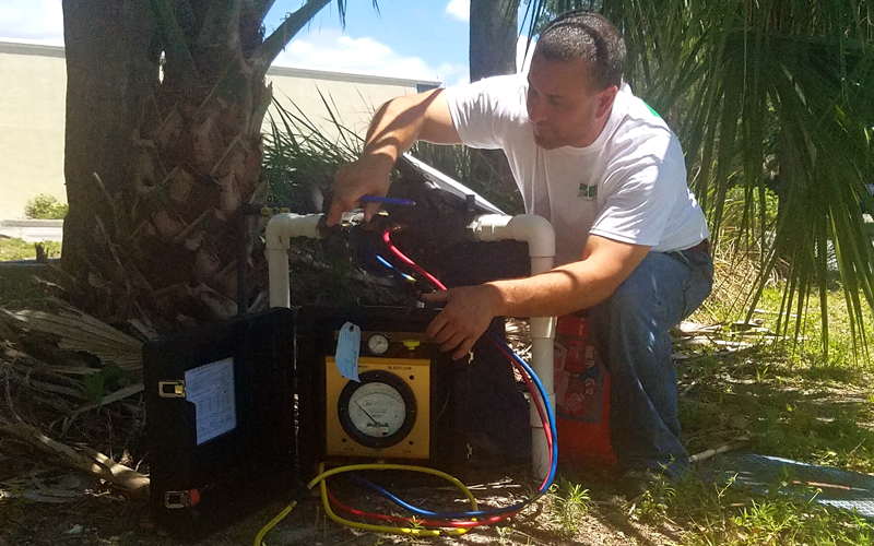 Man working on a black box with tubes, gauge, and wires outside under a tree.