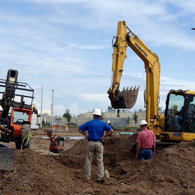 Construction site: workers in hard hats observing a yellow excavator digging in dirt under a cloudy sky.