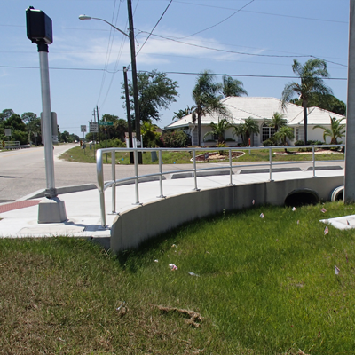Curb cut with railing, green grass, white building in background. Intersection with traffic signal on left.