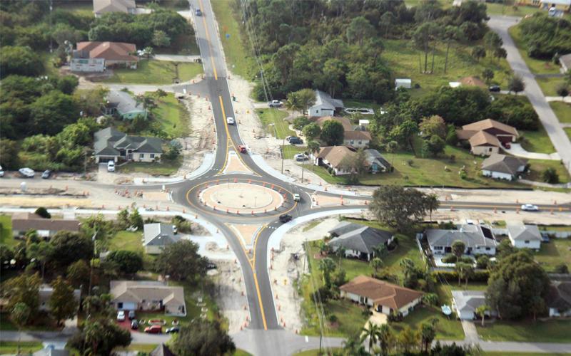 Aerial view of a roundabout under construction with several lanes, surrounded by houses and trees.