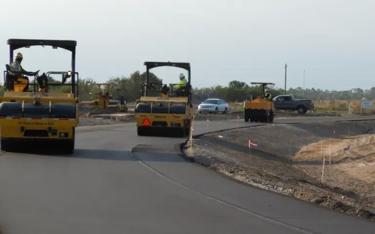 Road construction: three yellow asphalt rollers compacting fresh black asphalt on a curved road.
