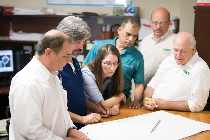 Team of six people reviewing architectural plans around a desk. Office setting, collaborative and focused.