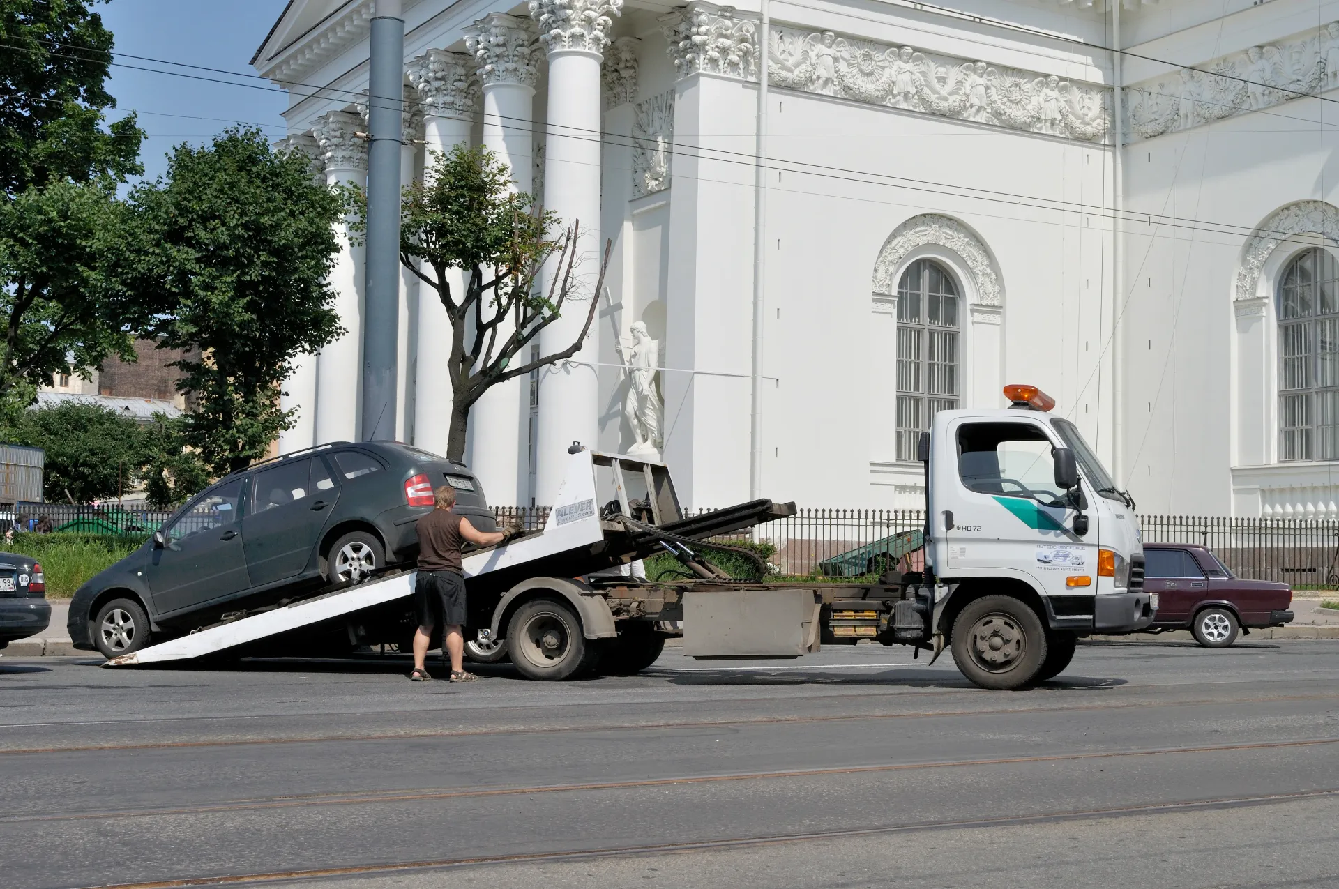 Tow truck loading a dark car on a city street in front of a white building.