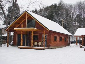 A log cabin in the snow with two chairs on the porch