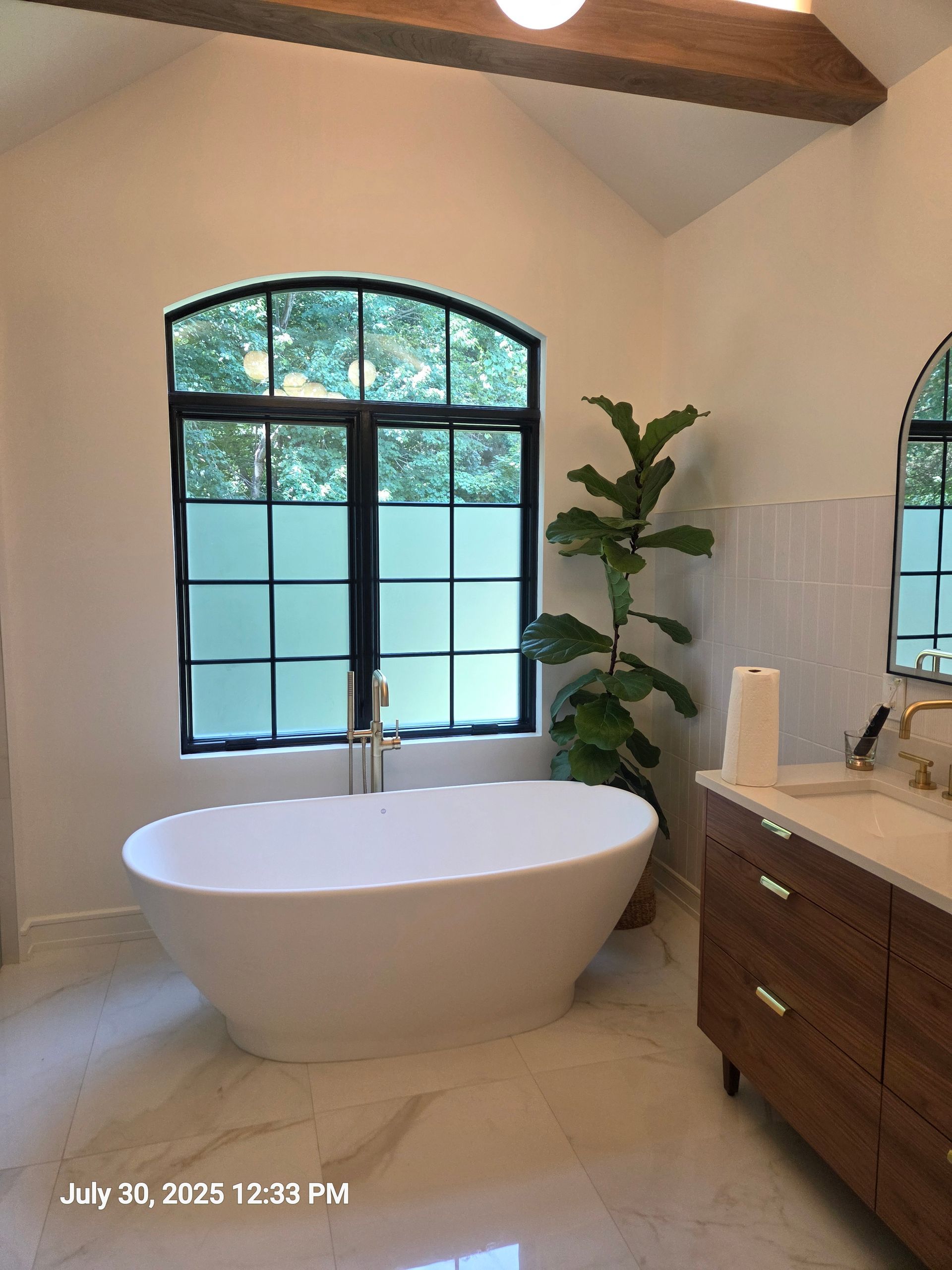 Modern bathroom with a freestanding tub, arched window, wood vanity, and plant accents
