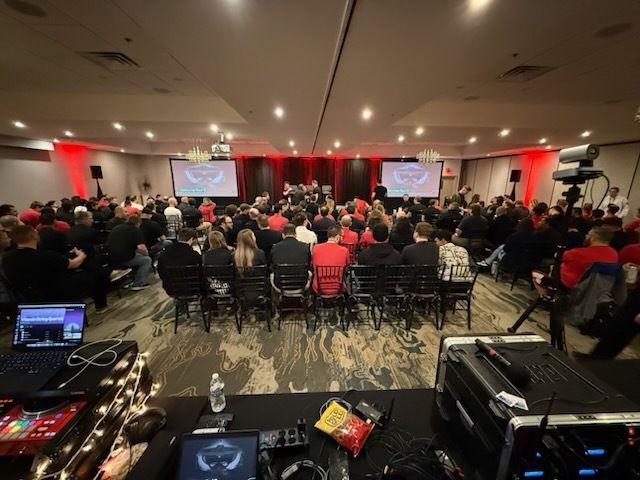 Conference audience seated in a ballroom facing two presentation screens and a stage with red drapes