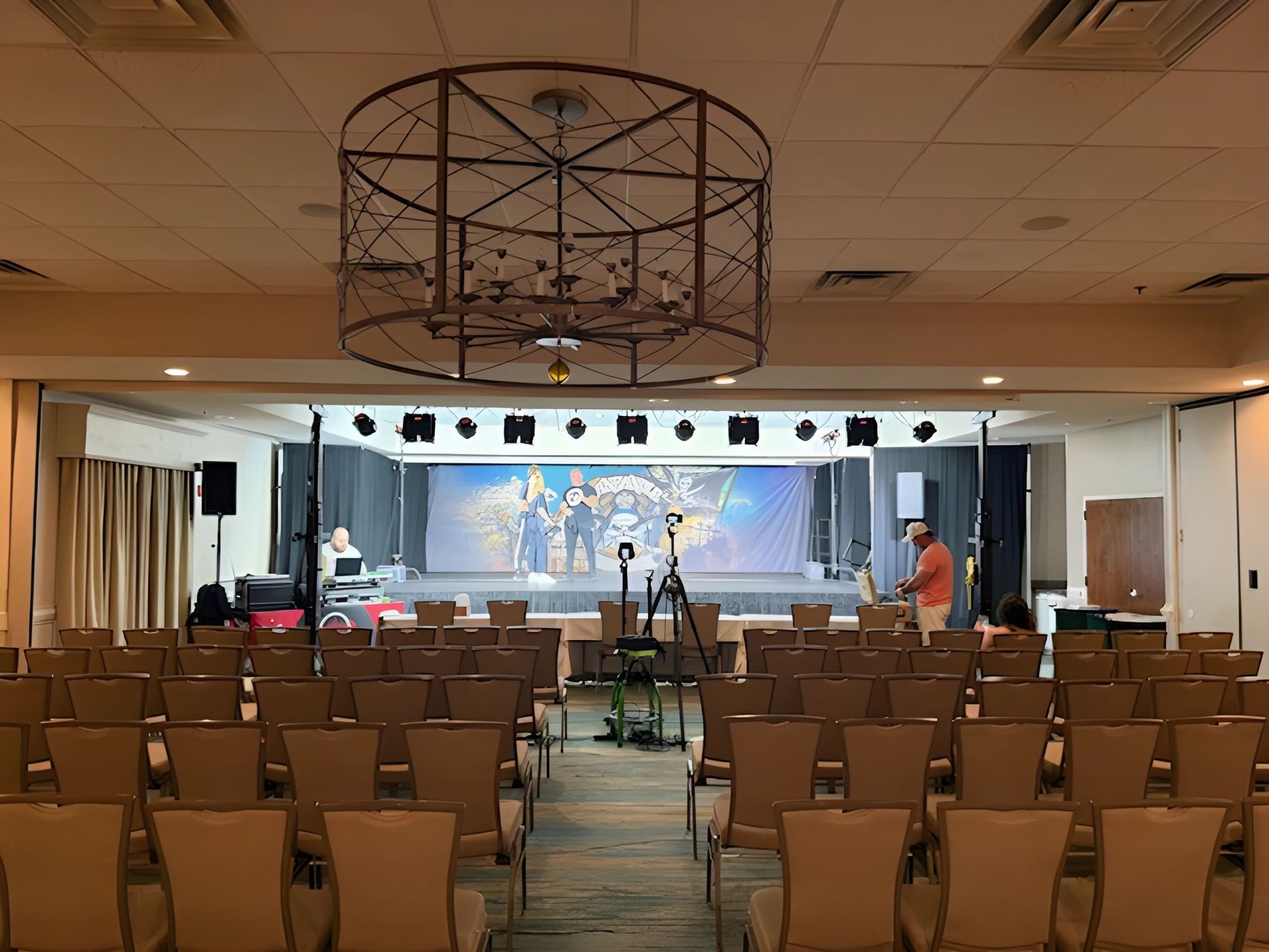 Empty conference room facing a lit stage with cameras, chairs, and a large screen at the front