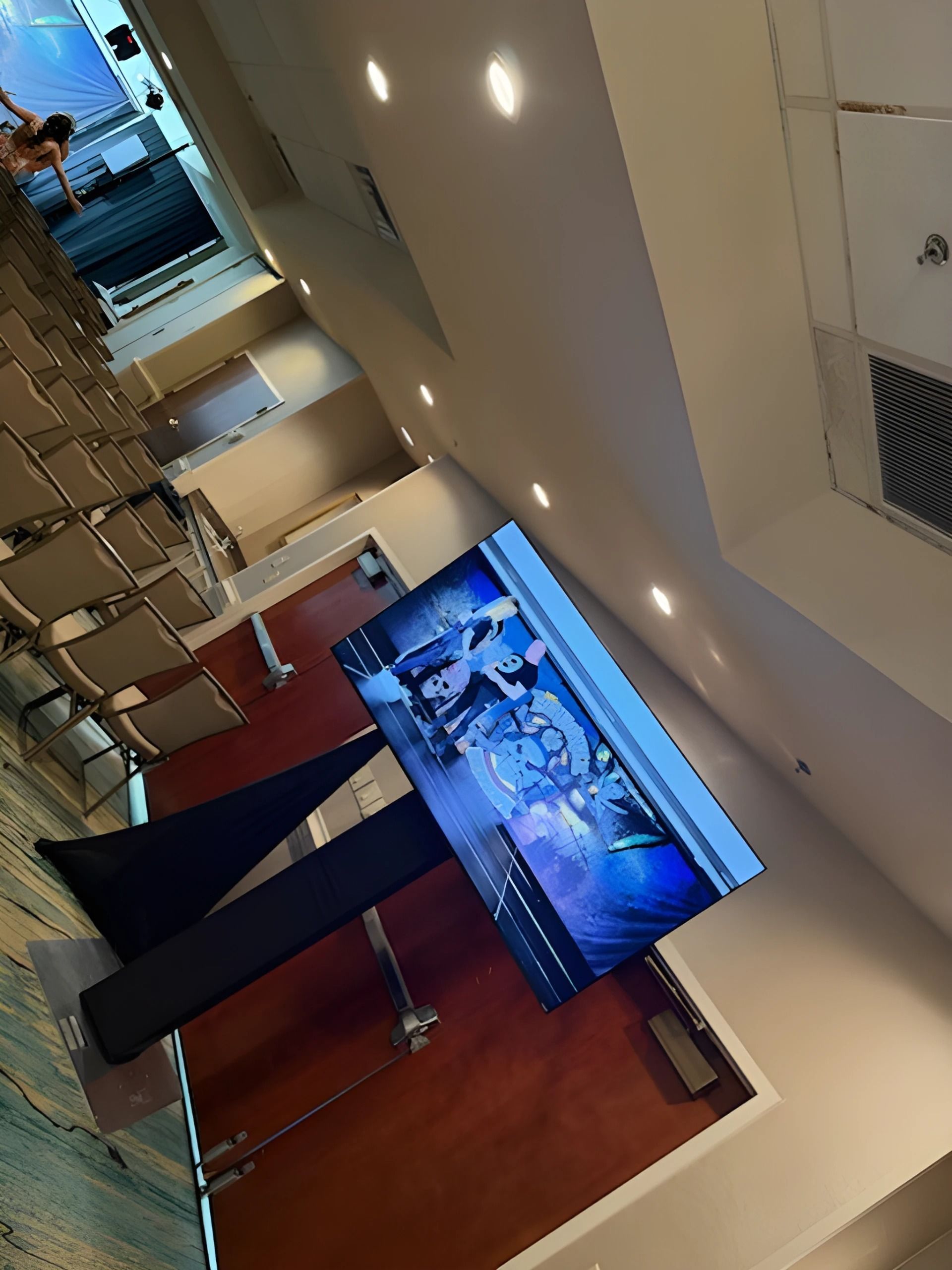 Conference room with rows of chairs, a podium, and a large screen showing a virtual meeting