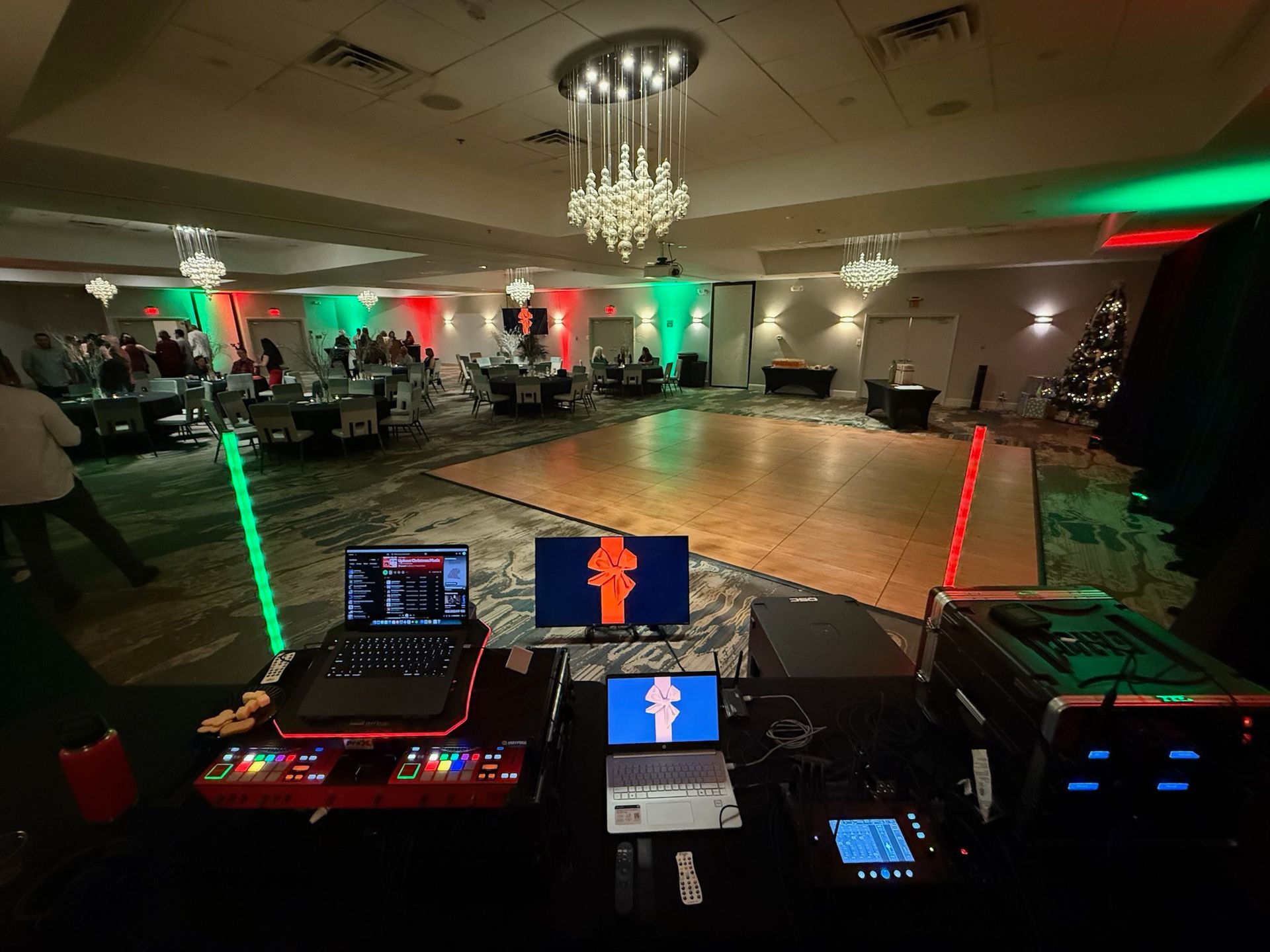 DJ booth facing a lit ballroom dance floor with guests and green/red uplights, chandeliers overhead