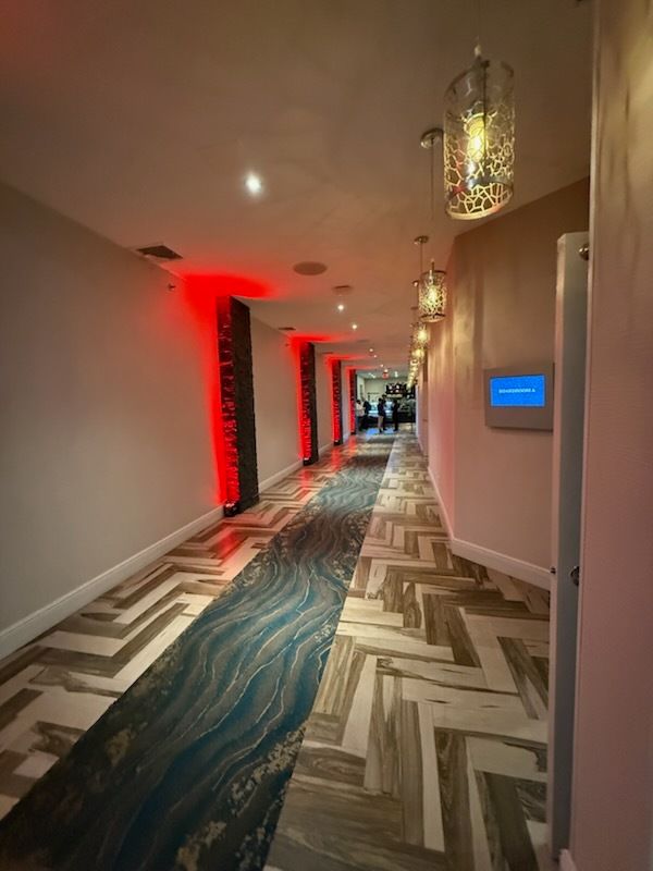 Dim hotel hallway with patterned carpet, red accent lighting, and ornate hanging lanterns.
