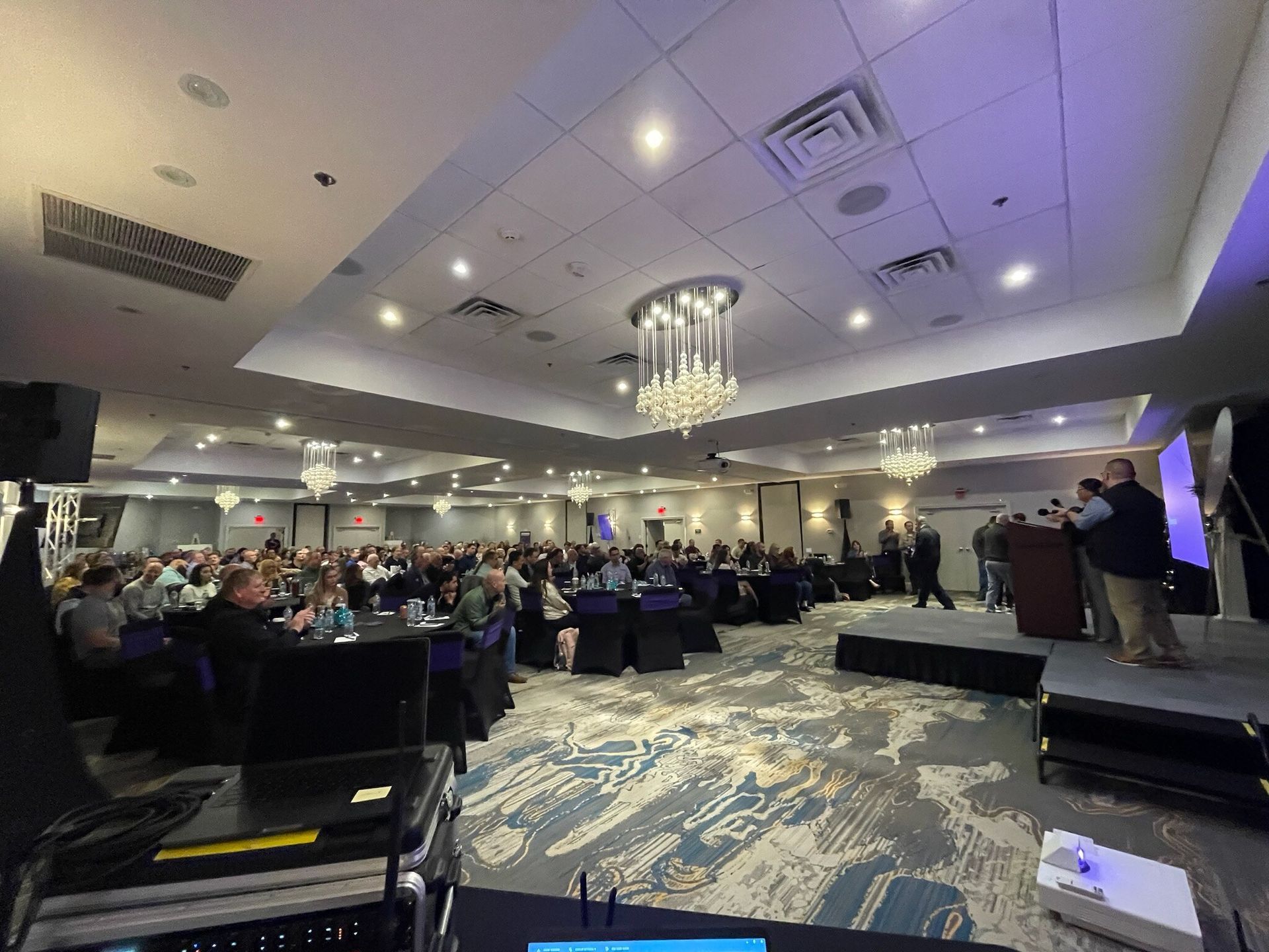 Banquet hall with guests seated at round tables, a speaker at a podium, and stage lighting overhead