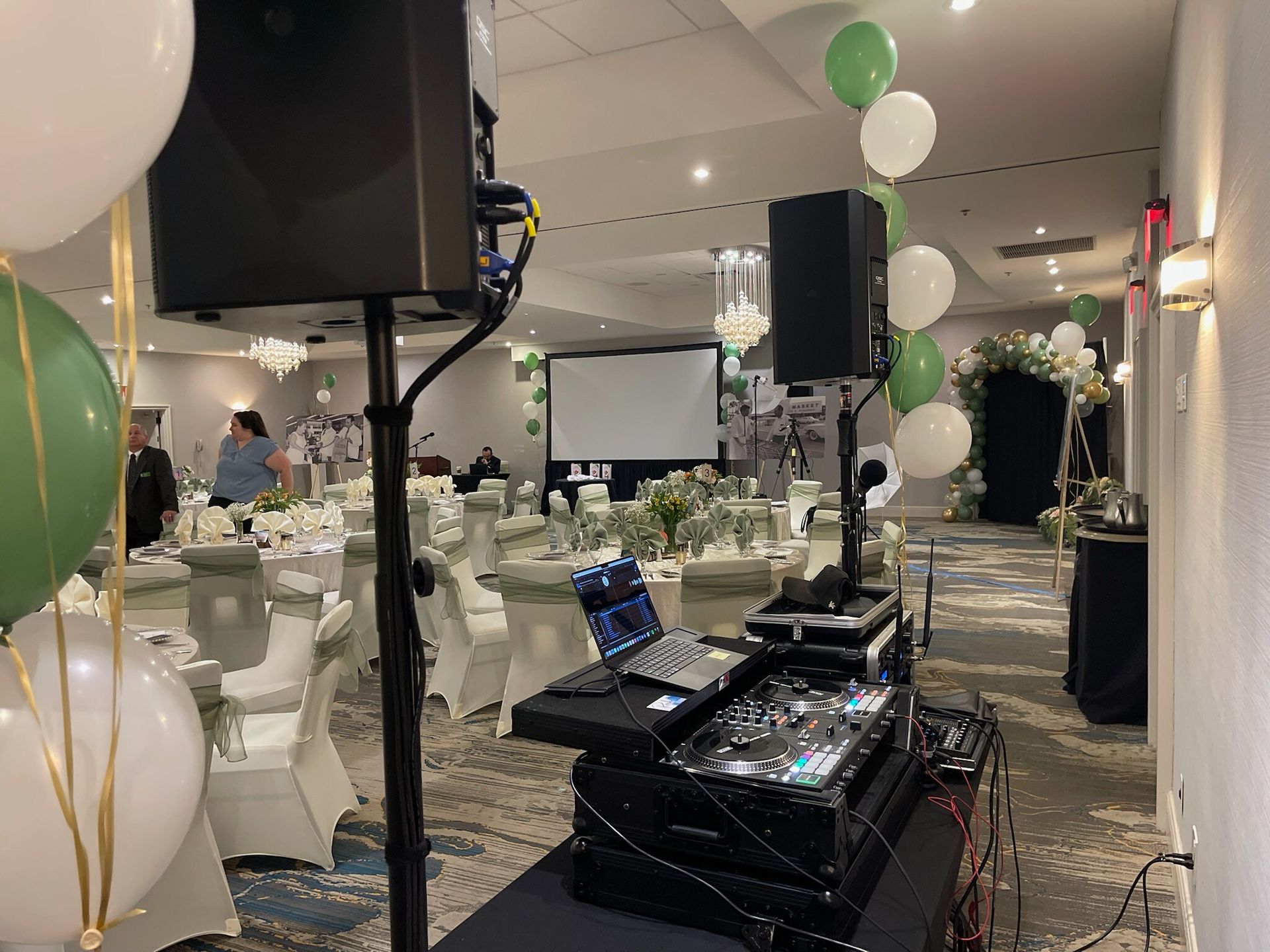 Decorated banquet hall with white and green balloons, tables, and a DJ setup in the foreground