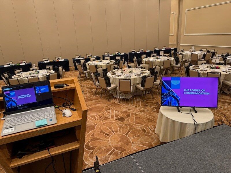Conference room with round banquet tables and twin monitors on a podium in the foreground.