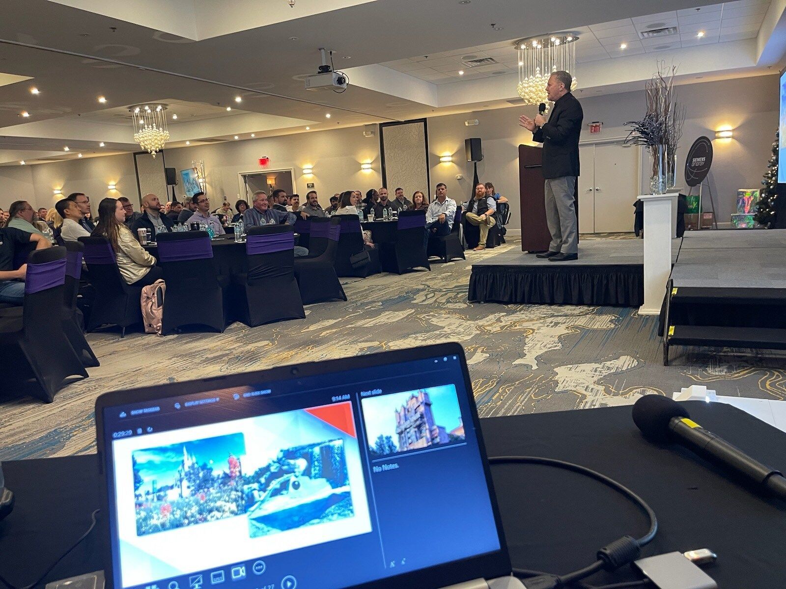 Presenter speaking on a stage at a conference, with an audience seated at round tables and a laptop in the foreground.