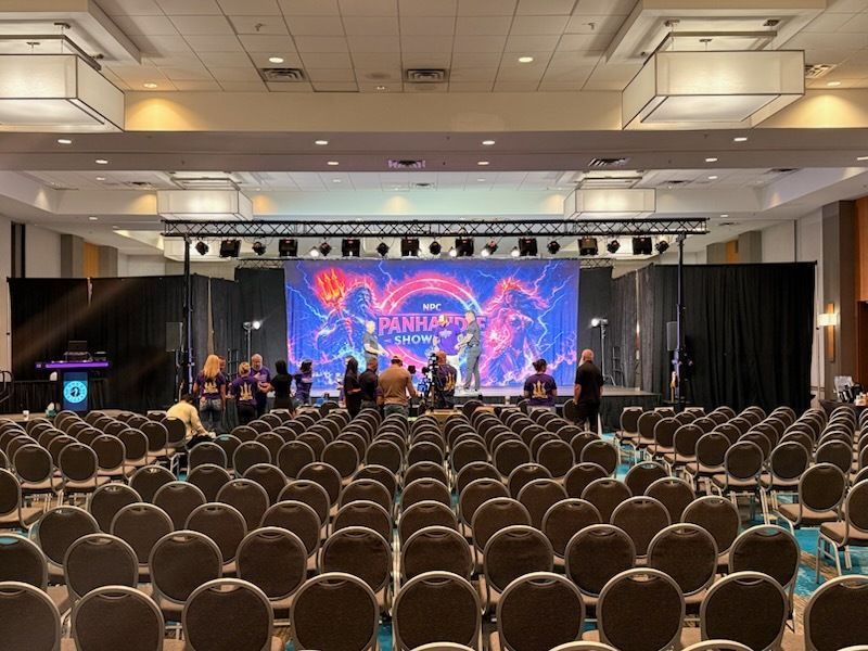 Conference hall with rows of chairs, a lit stage, and people setting up beneath a large colorful screen.