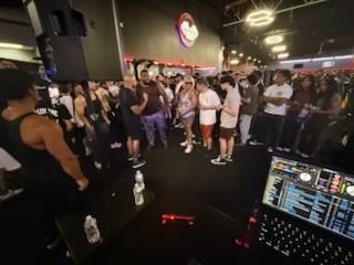 Crowded indoor dance class with people gathered on a dark floor near a DJ booth and stage lights