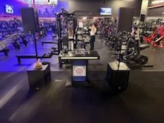 Gym floor with rows of exercise machines and purple lighting, with a person working out in the center.